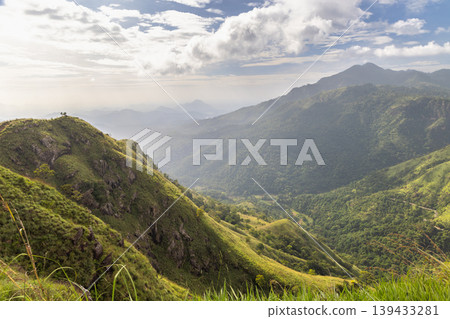 Dramatic Mountain Landscape View with Lush Green Slopes and Distant Peaks Under a Cloudy Sky at Golden Hour. View from Little Adam's Peak in Sri Lanka. Dramatic Mountain Landscape View with Lush Green Slopes and Distant Peaks Under a Cloudy Sky at Golden Hour. View from Little Adam's Peak in Sri Lanka. 139433281
