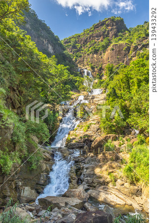 Ravana waterfall near Ella in Sri Lanka. Majestic multi-tiered waterfall cascades down a lush green mountain slope under a clear blue sky with white clouds, showcasing nature's beauty Ravana waterfall near Ella in Sri Lanka. Majestic multi-tiered waterfall cascades down a lush green mountain slope under a clear blue sky with white clouds, showcasing nature's beauty 139433292
