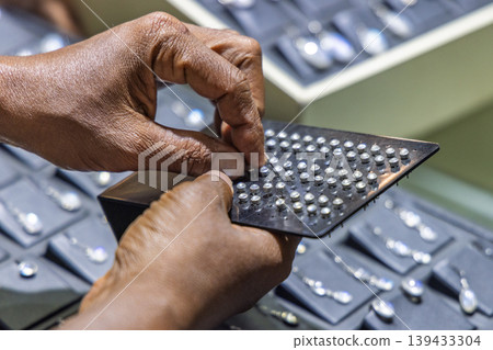 Close-up of a person's hands carefully selecting or arranging small, shimmering gem-like elements on a dark display tool. Sri Lanka island in Asia. 139433304