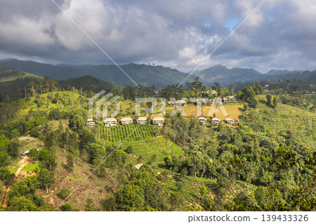 Panoramic view of rural hillside village nestled among lush green forests and terraced crops under cloudy sky. View from Little Adam's Peak in Sri Lanka. Panoramic view of rural hillside village nestled among lush green forests and terraced crops under cloudy sky. View from Little Adam's Peak in Sri Lanka. 139433326