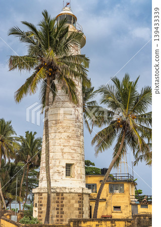 Historic white lighthouse tower stands tall amidst tropical palm trees under a bright blue sky, coastal architecture. Galle, the historic port city on the southwest coast of Sri Lanka. Historic white lighthouse tower stands tall amidst tropical palm trees under a bright blue sky, coastal architecture. Galle, the historic port city on the southwest coast of Sri Lanka. 139433339