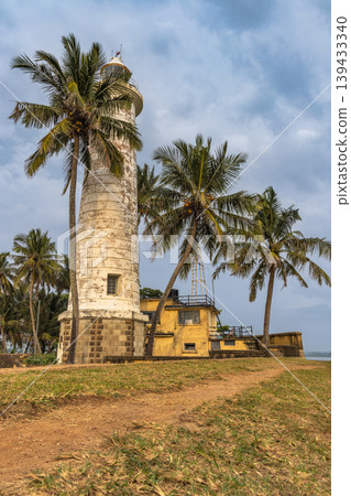 Historic Lighthouse with Palm Trees and Tropical Building on Coastal Landscape under Clouded Sky. Galle, the historic port city on the southwest coast of Sri Lanka. Historic Lighthouse with Palm Trees and Tropical Building on Coastal Landscape under Clouded Sky. Galle, the historic port city on the southwest coast of Sri Lanka. 139433340