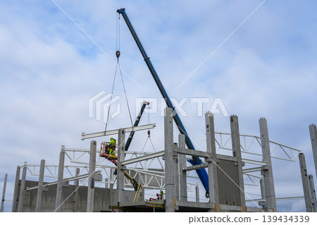 Crane lifting steel beam while worker on lift guides placement on concrete frame at build site 139434339