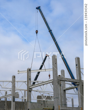 Crane hoists steel beam into place over concrete columns during industrial building construction 139434342