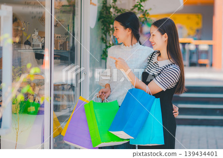 Two women are shopping in a store and pointing at the bags they are holding Two women are shopping in a store and pointing at the bags they are holding 139434401