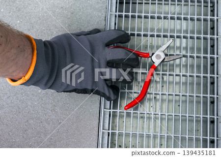 Close up of a hand in safety gloves adjusting a metal drain grate, highlighting practical outdoor repair and routine upkeep. Close up of a hand in safety gloves adjusting a metal drain grate, highlighting practical outdoor repair and routine upkeep. 139435181