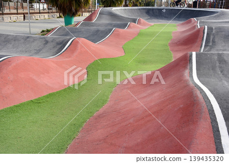 Empty coastal pump track with undulating asphalt waves and colorful lanes near marina for biking and skate sports Empty coastal pump track with undulating asphalt waves and colorful lanes near marina for biking and skate sports 139435320