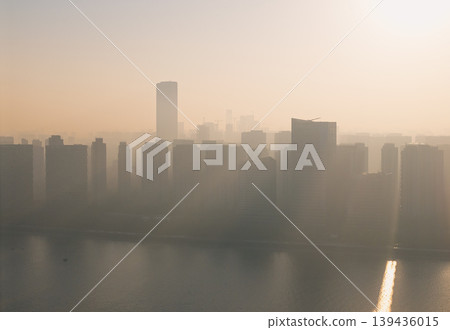 Aerial views of a Chinese cityscape at sunrise show buildings rising above a river. Early morning sun rays shine through the city haze. Aerial views of a Chinese cityscape at sunrise show buildings rising above a river. Early morning sun rays shine through the city haze. 139436015