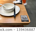 QR code on wooden restaurant table for digital menu and scan to order service. Modern contactless dining technology with white ceramic bowl and chopsticks in blurred background setting 139436887