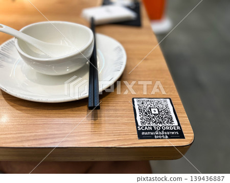 QR code on wooden restaurant table for digital menu and scan to order service. Modern contactless dining technology with white ceramic bowl and chopsticks in blurred background setting QR code on wooden restaurant table for digital menu and scan to order service. Modern contactless dining technology with white ceramic bowl and chopsticks in blurred background setting 139436887