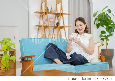 A middle-aged Japanese woman is sitting on the living room sofa and using her smartphone. A middle-aged Japanese woman is sitting on the living room sofa and using her smartphone. 139437508