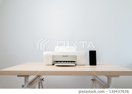 Minimal workspace featuring a white printer and a black smartphone on a light wooden desk against a neutral wall, evoking calm, simplicity, technology, and small home office or studio setup. Minimal workspace featuring a white printer and a black smartphone on a light wooden desk against a neutral wall, evoking calm, simplicity, technology, and small home office or studio setup. 139437636