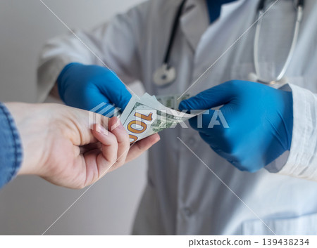 Close-up of doctor in white coat and blue gloves receiving or hiding cash. Concept of high medical salary, corruption in medicine, healthcare bribery, and expensive health insurance. Close-up of doctor in white coat and blue gloves receiving or hiding cash. Concept of high medical salary, corruption in medicine, healthcare bribery, and expensive health insurance. 139438234