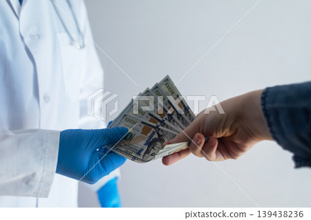 Close-up of doctor in white coat and blue gloves receiving or hiding cash. Concept of high medical salary, corruption in medicine, healthcare bribery, and expensive health insurance. Close-up of doctor in white coat and blue gloves receiving or hiding cash. Concept of high medical salary, corruption in medicine, healthcare bribery, and expensive health insurance. 139438236