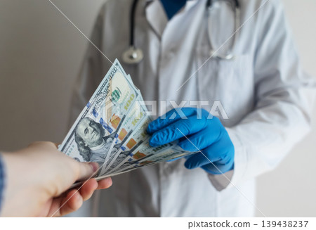 Close-up of doctor in white coat and blue gloves receiving or hiding cash. Concept of high medical salary, corruption in medicine, healthcare bribery, and expensive health insurance. 139438237