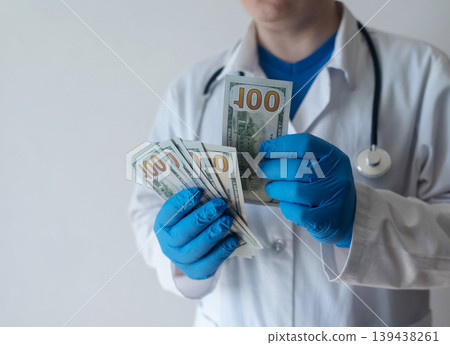Close-up of doctor in white coat and blue gloves receiving or hiding cash. Concept of high medical salary, corruption in medicine, healthcare bribery, and expensive health insurance. 139438261