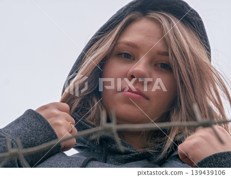 Young woman with an intense gaze in a dark hoodie behind a barbed wire fence on an overcast day 139439106