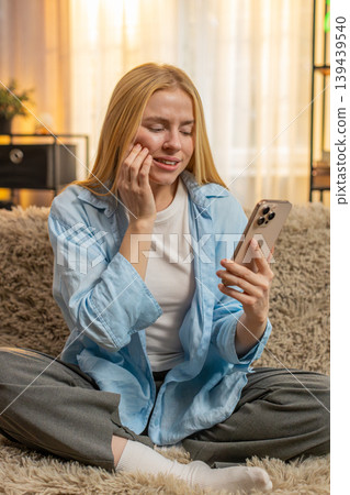 Young woman feeling tooth pain at home, worried face, searching phone for dentist, sofa discomfort 139439540