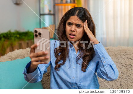 Indian woman looking down with sad sigh while reading bad news on smartphone on home sofa indoors Indian woman looking down with sad sigh while reading bad news on smartphone on home sofa indoors 139439570