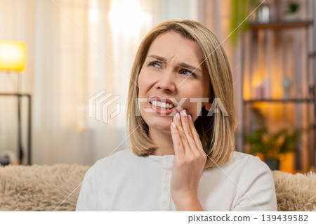 Adult woman with toothache at home, holding jaw and showing pain expression while sitting on sofa Adult woman with toothache at home, holding jaw and showing pain expression while sitting on sofa 139439582