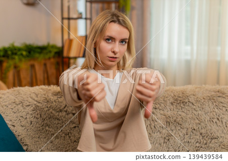 Woman showing thumbs down gesture at home, expressing clear dissatisfaction with serious expression Woman showing thumbs down gesture at home, expressing clear dissatisfaction with serious expression 139439584