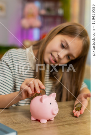 Child girl kid happily saves coins into piggy bank smiling at table on home sofa planning new toy Child girl kid happily saves coins into piggy bank smiling at table on home sofa planning new toy 139439589