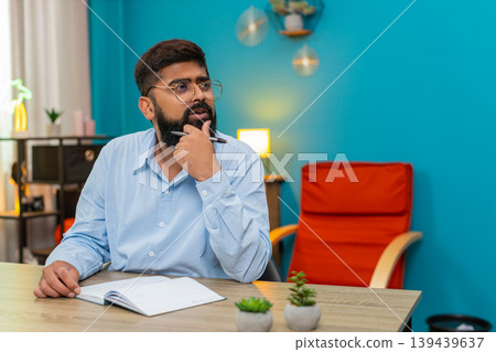 Indian man holds pen gazes notebook deeply thinking planning ideas writing strategy indoors focused Indian man holds pen gazes notebook deeply thinking planning ideas writing strategy indoors focused 139439637