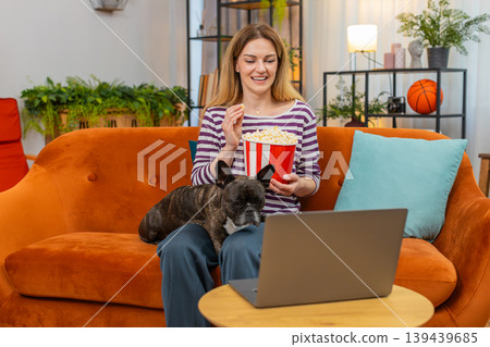 Middle-aged woman with dog laughs eats popcorn and watches favorite series or movie on laptop calm Middle-aged woman with dog laughs eats popcorn and watches favorite series or movie on laptop calm 139439685