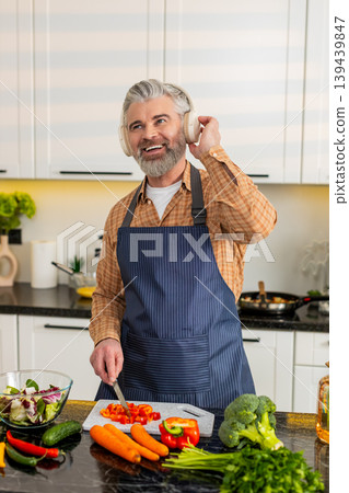 Middle-aged man chopping red pepper wearing headphones enjoying music while preparing healthy meal Middle-aged man chopping red pepper wearing headphones enjoying music while preparing healthy meal 139439847