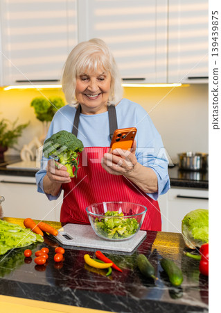 Senior old woman checking smartphone while making healthy vegetable salad in home kitchen diet plan 139439875