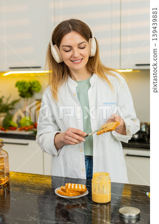 Middle-aged woman dancing with headphones in home kitchen singing while making healthy diet toast 139439881
