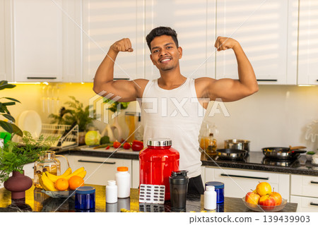 Indian man showing strong biceps near protein supplements in kitchen for fitness nutrition routine Indian man showing strong biceps near protein supplements in kitchen for fitness nutrition routine 139439903