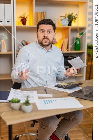 Young man on home table checking bills and reacting to unexpected payments with frustration stress Young man on home table checking bills and reacting to unexpected payments with frustration stress 139439911