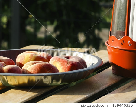 Flat peaches in a metal bowl and red lantern on a wooden table, creating a seasonal fruit still life with a rustic natural setting Flat peaches in a metal bowl and red lantern on a wooden table, creating a seasonal fruit still life with a rustic natural setting 139440330