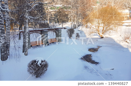 Snow-covered park scene featuring a stone bench structure with wooden seating, surrounded by trees and a small stream in a winter landscape 139440827