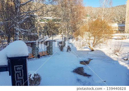 Snow-covered winter landscape featuring a frozen stream, bare trees, and a stone bench in a serene outdoor setting surrounded by snow-laden hills 139440828