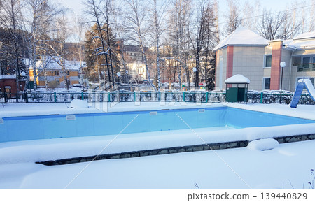 Outdoor swimming pool covered in snow during winter season, surrounded by trees and buildings, with a clear blue sky above and a playground visible in the background 139440829