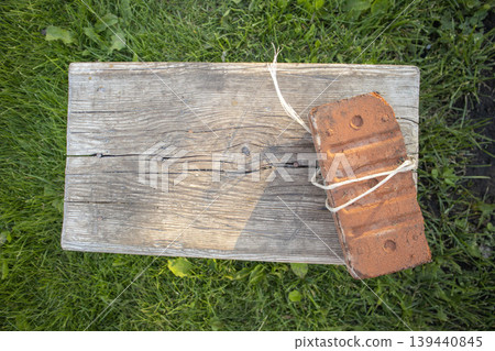 Wooden plank with a tied red brick resting on top, surrounded by green grass in a natural outdoor setting under soft sunlight Wooden plank with a tied red brick resting on top, surrounded by green grass in a natural outdoor setting under soft sunlight 139440845