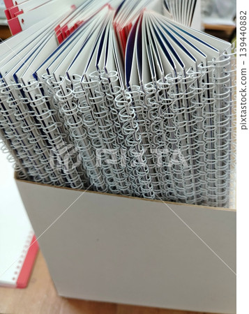 Stack of spiral-bound notebooks with colorful covers arranged neatly in a white storage box on a wooden surface in an office or study environment 139440882