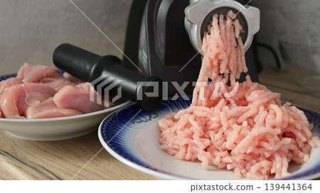 A domestic meat grinder grinds minced meat from a meat fillet in the kitchen, located on a light countertop against a textured gray wall, the operation of kitchen appliances A domestic meat grinder grinds minced meat from a meat fillet in the kitchen, located on a light countertop against a textured gray wall, the operation of kitchen appliances 139441364