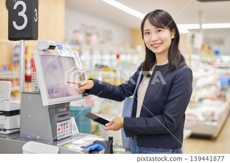A young woman using her smartphone to pay at a self-checkout counter. 139441877