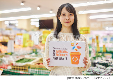 A young woman holding a sign about food waste in a supermarket aisle. A young woman holding a sign about food waste in a supermarket aisle. 139442275