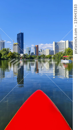Red paddleboard on water with modern city skyline reflection. Summer water sport, mental vitality, burst of energy. Red paddleboard on water with modern city skyline reflection. Summer water sport, mental vitality, burst of energy. 139443583