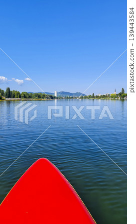Red paddleboard in the middle of a calm river under a clear blue sky. Summer water sport, mental vitality, burst of energy. 139443584