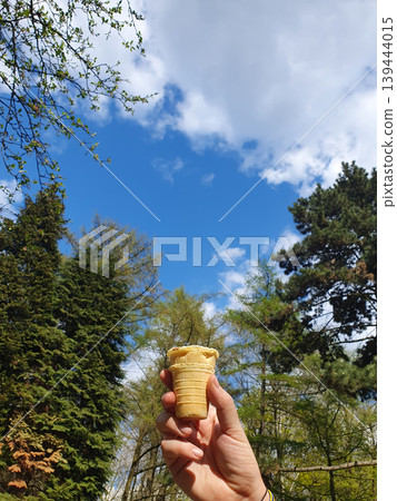 Hand holding a waffle cup of ice cream against a bright blue sky and green trees. Concept of summer vacation Hand holding a waffle cup of ice cream against a bright blue sky and green trees. Concept of summer vacation 139444015