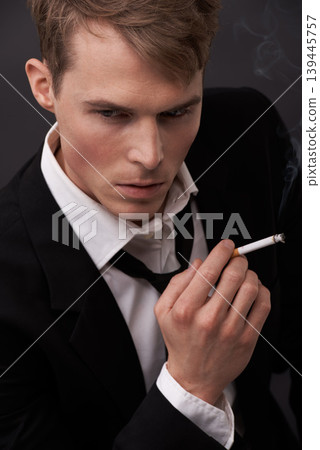 Face, thinking and a man smoking a cigarette in studio on a dark background for tobacco addiction. Nicotine, habit and unhealthy with a young person in a suit, taking a break from business to smoke Face, thinking and a man smoking a cigarette in studio on a dark background for tobacco addiction. Nicotine, habit and unhealthy with a young person in a suit, taking a break from business to smoke 139445757
