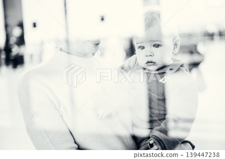 Thoughtful young mother looking trough window holding his infant baby boy child while waiting to board an airplane at airport terminal departure gates. Travel with baby concept 139447238
