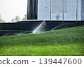 Automatic sprinkling system watering green grass in city park. Lawn irrigation device spraying water drops on meadow field near modern building architecture. Care of urban landscape. 139447600