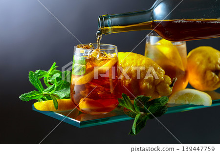 Black tea is poured from a bottle into a glass with lemon wedges, ice cubes and mint leaves. Black tea is poured from a bottle into a glass with lemon wedges, ice cubes and mint leaves. 139447759
