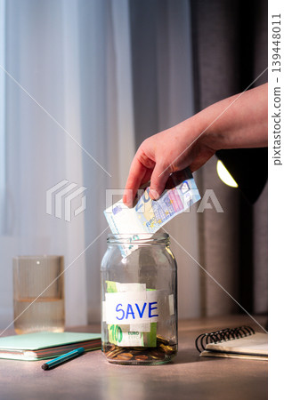 Close up of cash being added to transparent money jar. Represents traditional saving methods and conscious money management for future security. Vertical image Close up of cash being added to transparent money jar. Represents traditional saving methods and conscious money management for future security. Vertical image 139448011